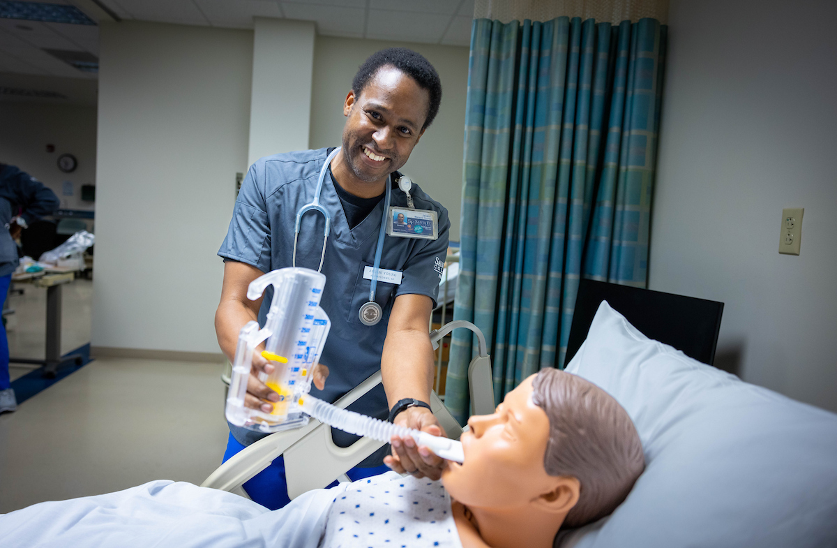 An individual holds a device to a mannequin's mouth in a medical setting.