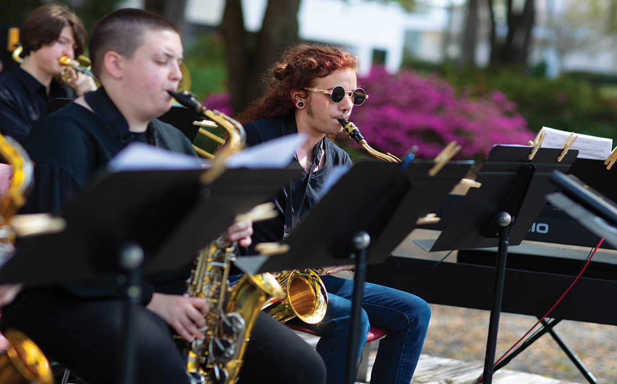 Three musicians perform on an outdoor stage, each playing their instrument.