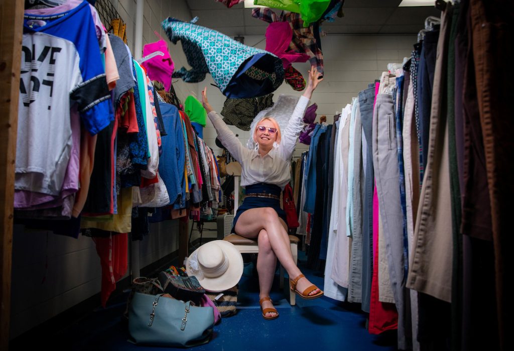  A person sitting with arms raised, surrounded by racks of clothes on both sides.