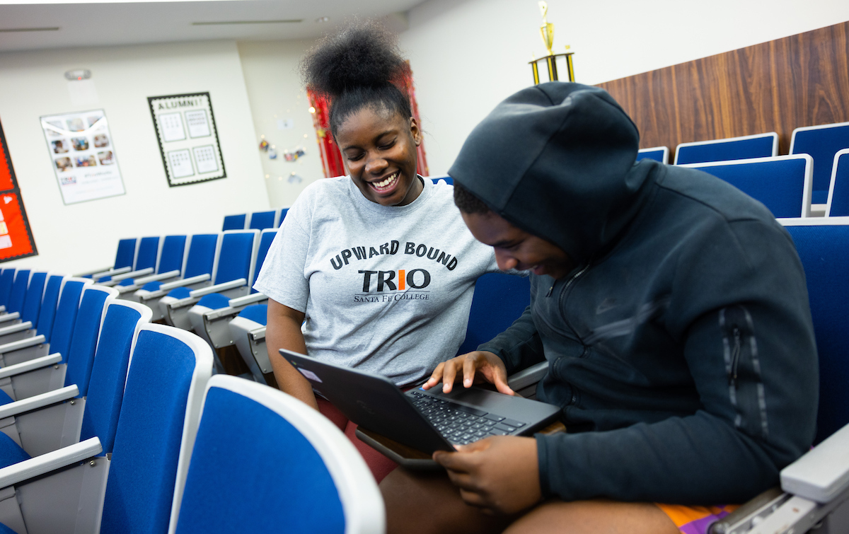 Two individuals smiling and looking at a laptop, one wearing a shirt that says 