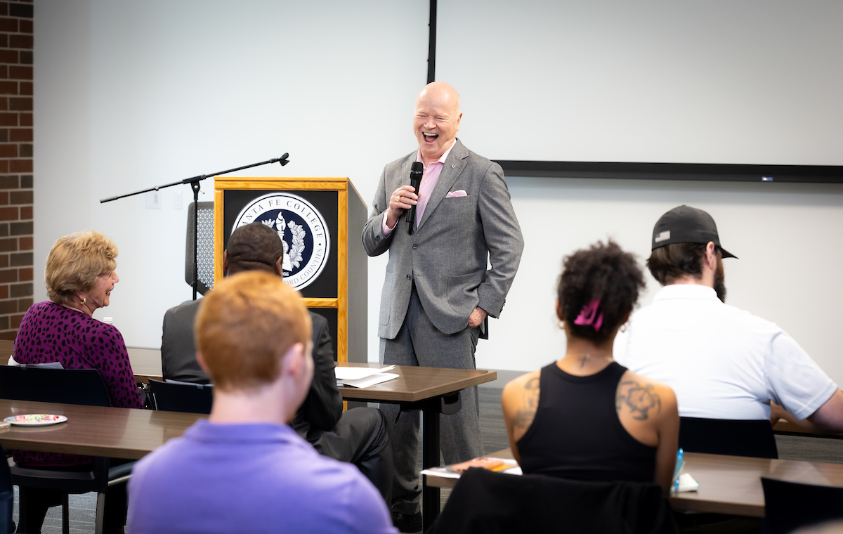 An individual laughs while presenting in front of a seated audience in a meeting room.