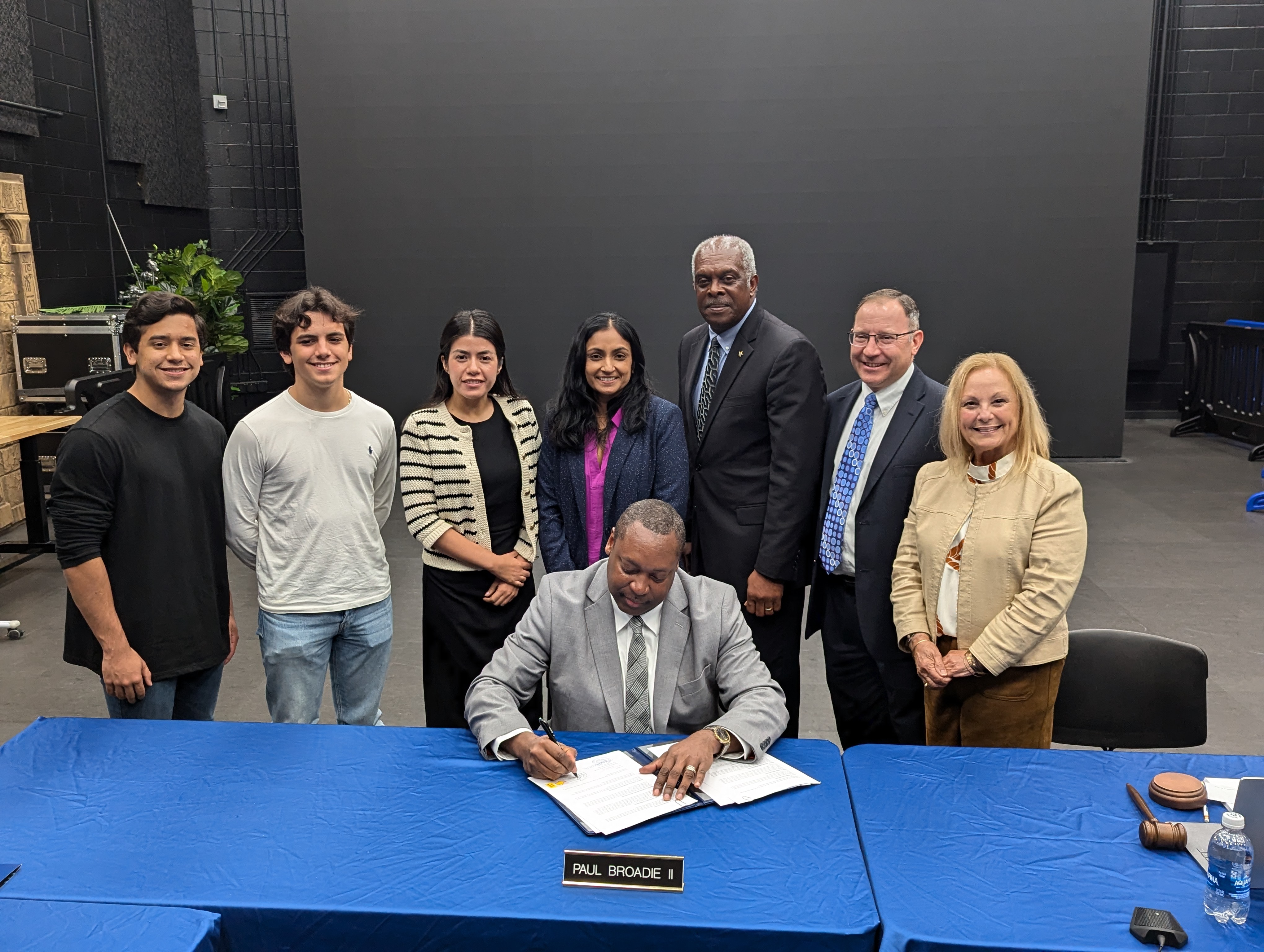 Dr. Broadie surrounded by students, staff and board members signs document