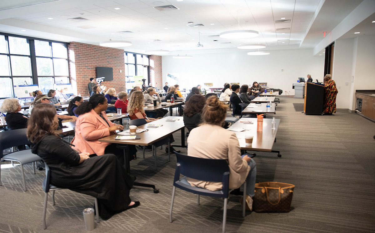 A diverse group of attendees seated at tables, attentively listening to a speaker at a symposium in a well-lit room.