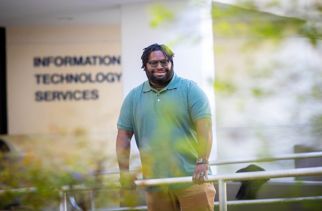 Environmental portrait of Dominic Perry, Santa Fe College A.S. Information Technology graduate stands in front of the ITE building wearing a green shirt. 