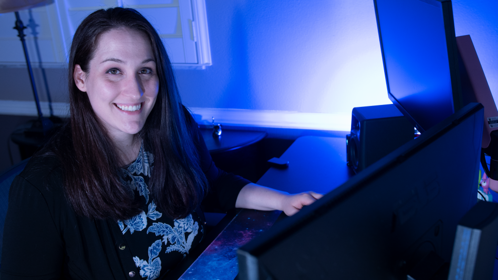 Santa Fe College graduate sits in front of a computer at a desk, illuminated in blue light smiling at the camera wearing a floral shirt and black jacket. 