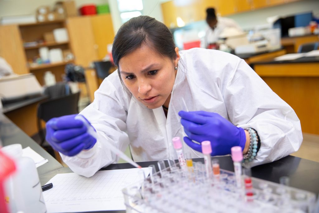 A student dressed in a white lab coat and blue gloves places a liquid into a test tube via a dropper.