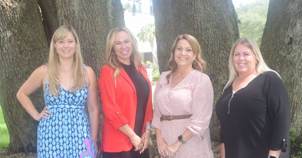 A group of four women poses gracefully, smiling, while standing united in front of a majestic tree.