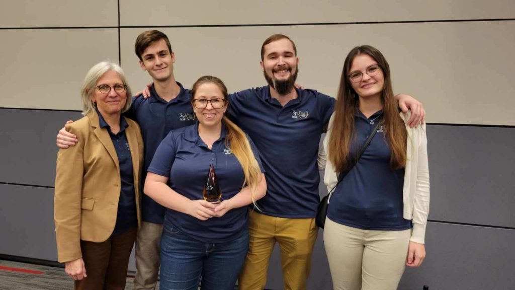 Five people in blue shirts standing together, one person proudly holds an award in his hand.