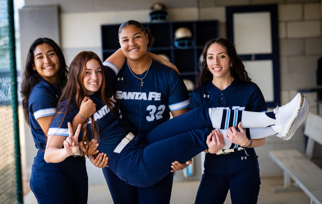 Santa Fe College Saints Softball players Ariel Obregon, Alexis Daphney, Anissa Quinonez and Sophia Davis in a dugout.